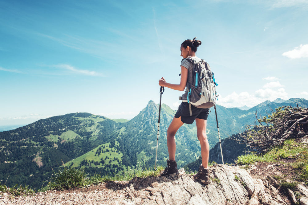 A woman hiking stands atop a ridge and overlooks an expansive, alpine valley.